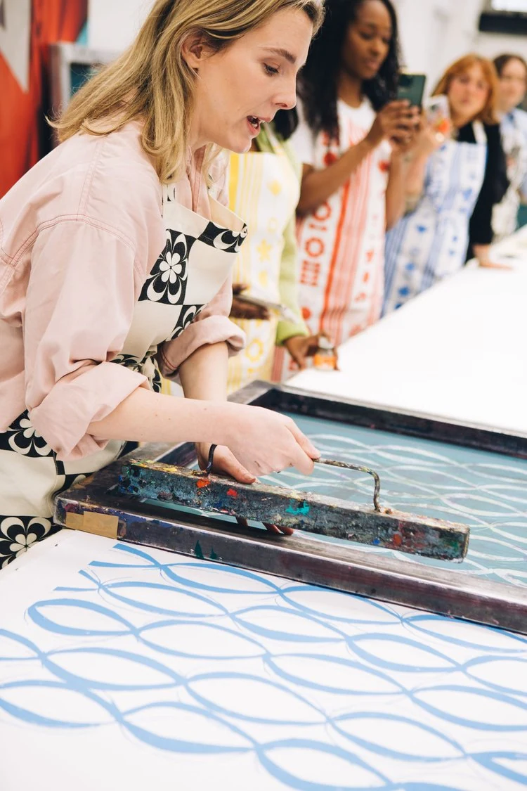 A woman in a pink shirt and patterned apron presses a squeegee on a large piece of paper with blue geometric designs, while four women in colorful aprons watch and take photos in the background.