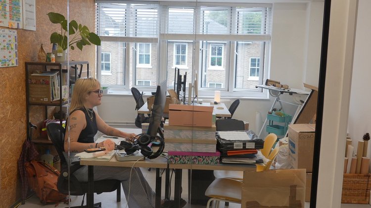 An office with a woman working at a desk, facing computer monitors, cluttered with documents, boxes, and office supplies.