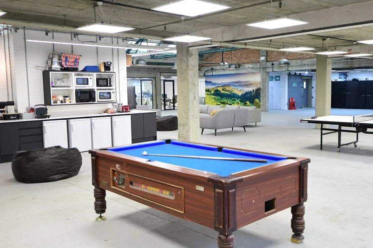 Indoor recreational lounge with a pool table, bean bag chair, and kitchenette area featuring microwaves and a toaster. There is a sofa and a landscape mural on the wall, with glass doors leading to another room, all within an industrial-style space with exposed concrete and ceiling pipes.