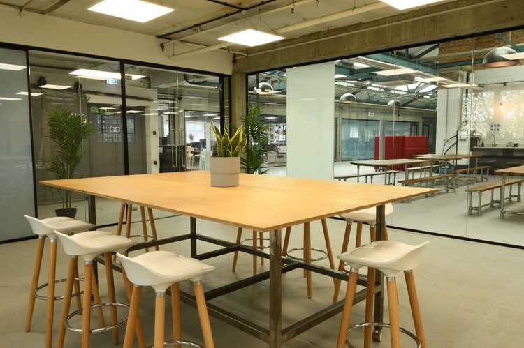 Modern industrial office kitchen or dining area with a wooden table, white barstools, potted plants, and glass walls revealing additional seating and workspaces in the background.