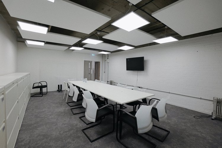 Empty conference room with a long white table, white and black chairs, a wall-mounted television, and ceiling lighting panels.