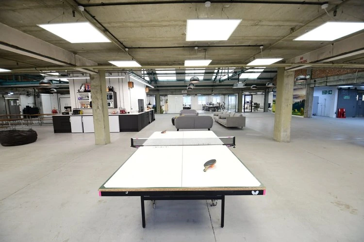Indoor space with a ping pong table in the foreground and couches and a kitchenette in the background, suggesting a lounge area in an office or recreational facility.