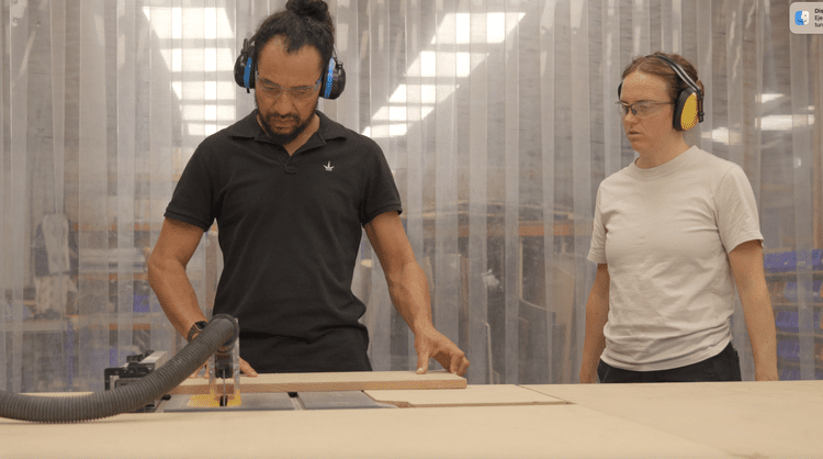 Two people wearing safety glasses and ear protection in a woodworking shop, one operating a woodworking machine and the other observing.