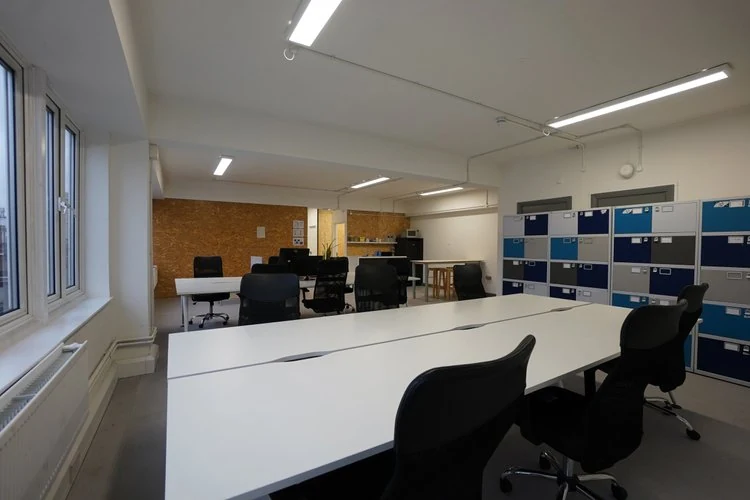 Shared office desks and chairs beside windows and blue-and-white lockers.