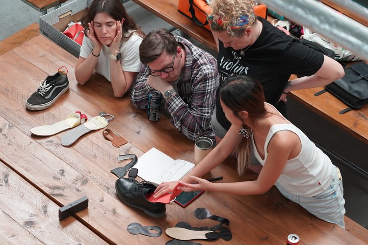 Four young women and one young man gathered around a wooden table looking at various shoe insoles and shoe related items, with one woman showing a shoe insert.