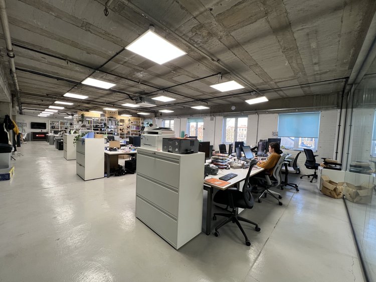 Open office with desks, computers and people working beneath a concrete ceiling.