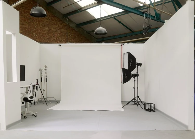 Photography studio setup with white backdrop, lighting equipment, tripod, chair, and computer in a warehouse with a brick wall and metal roof.