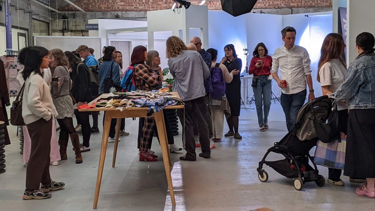 A group of people shopping at an indoor market or craft fair, with tables of fabric and clothing, some wearing backpacks and pushing strollers.