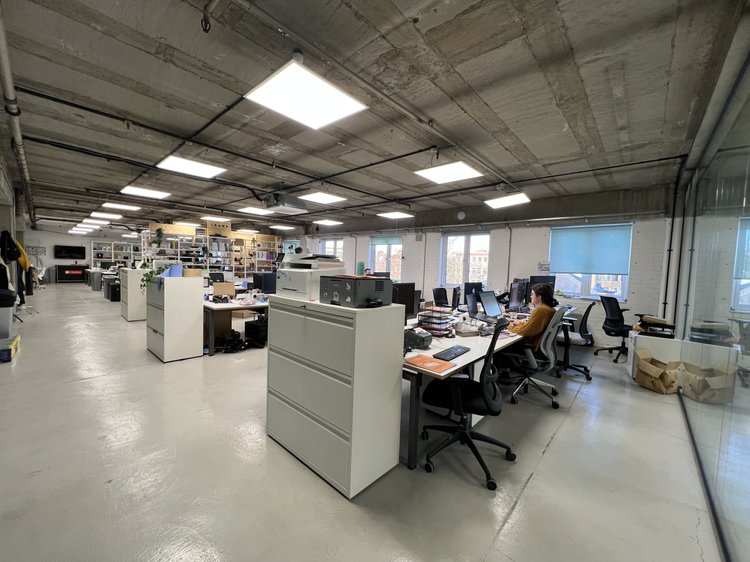 Open office with desks, computers and people working beneath a concrete ceiling.