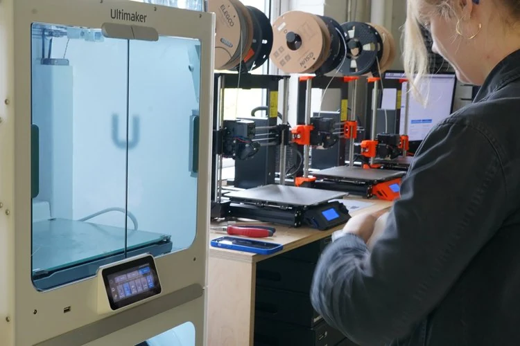 A person working with 3D printers in a workshop, surrounded by spools of filament and 3D printing equipment.